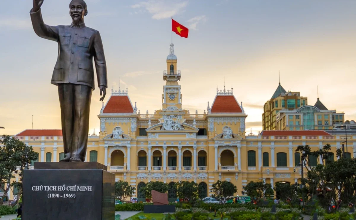 A large bronze statue of Ho Chi Minh, with the inscription in Vietnamese and years 1890-1969, standing in front of a historic yellow building with red roof towers, a clock tower, and the Vietnamese flag flying on top, in a cityscape during sunset.