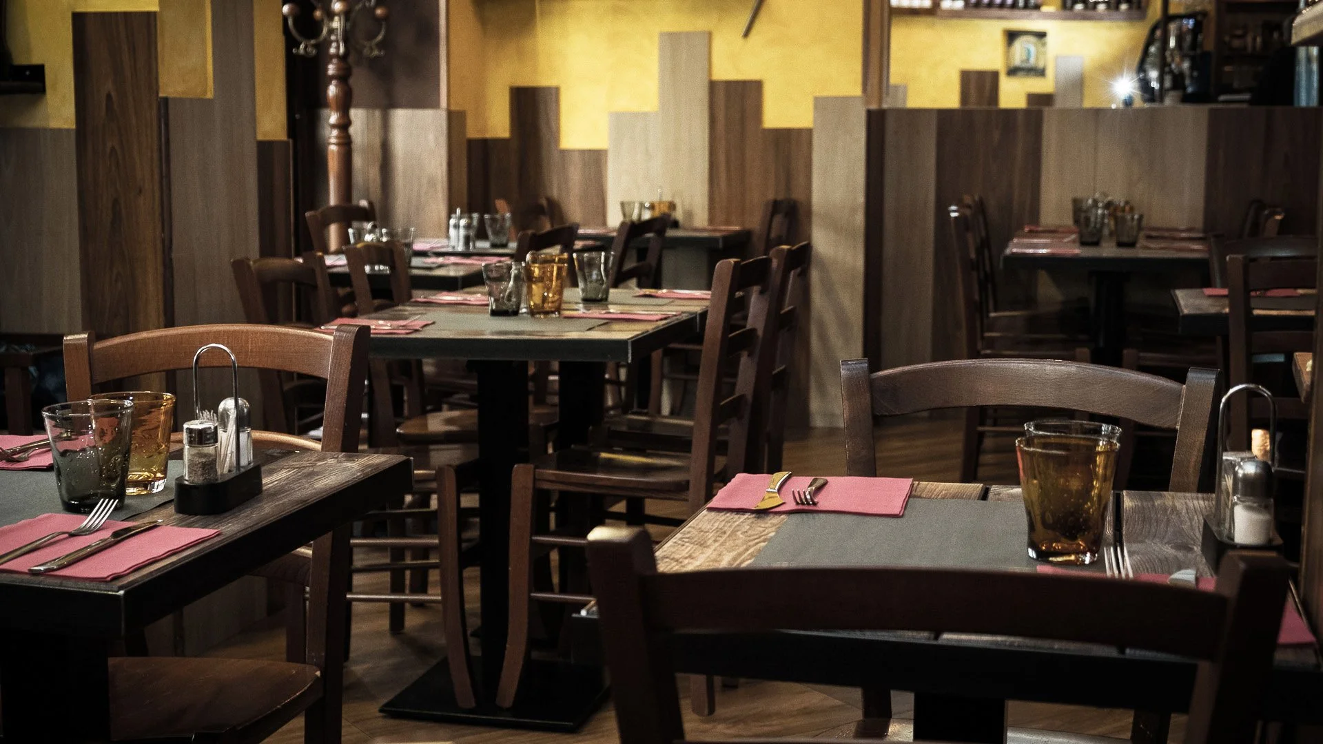 Empty restaurant dining area with wooden chairs, tables set with glasses, napkins, and cutlery, and a warm yellow and brown wall in the background.