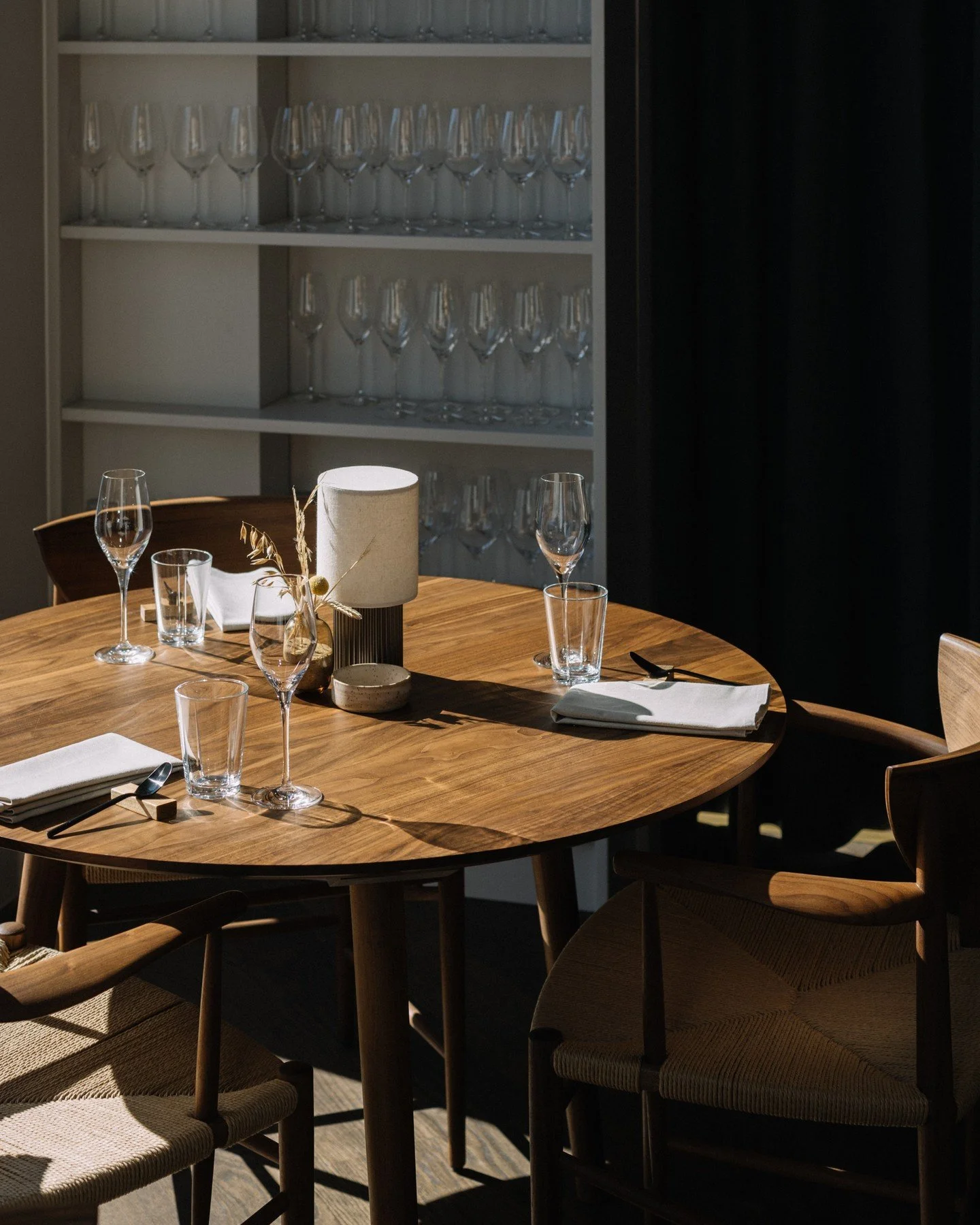 A round wooden dining table set with wine glasses, napkins, and a centerpiece lamp in a dimly lit room with a shelf of wine glasses in the background.