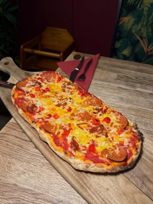 A long, thin pizza with pepperoni, sausage, melted cheese, and red peppers on a wooden serving board.