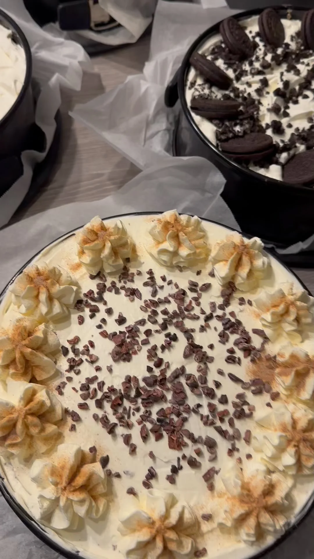 Two large cakes decorated with whipped cream, chocolate shavings, and Oreo cookies, placed on a table with parchment paper.