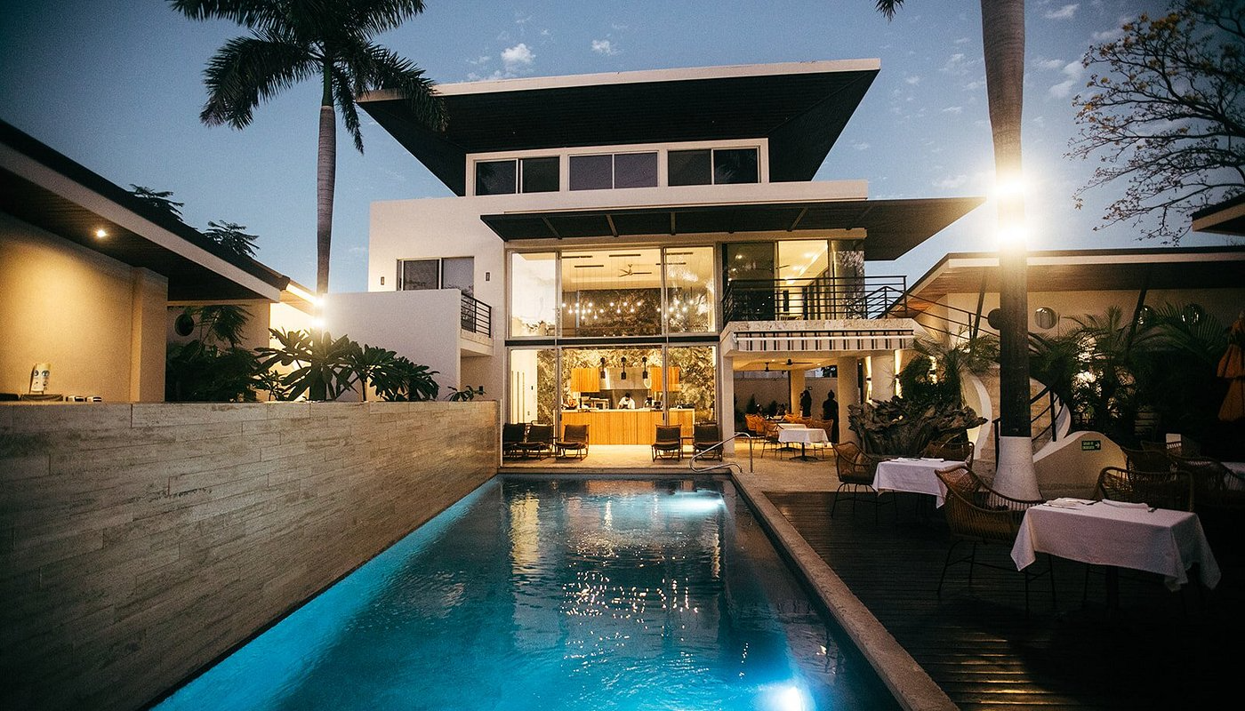 A modern house with a swimming pool outside, dining tables with white tablecloths, and tropical plants, during sunset.
