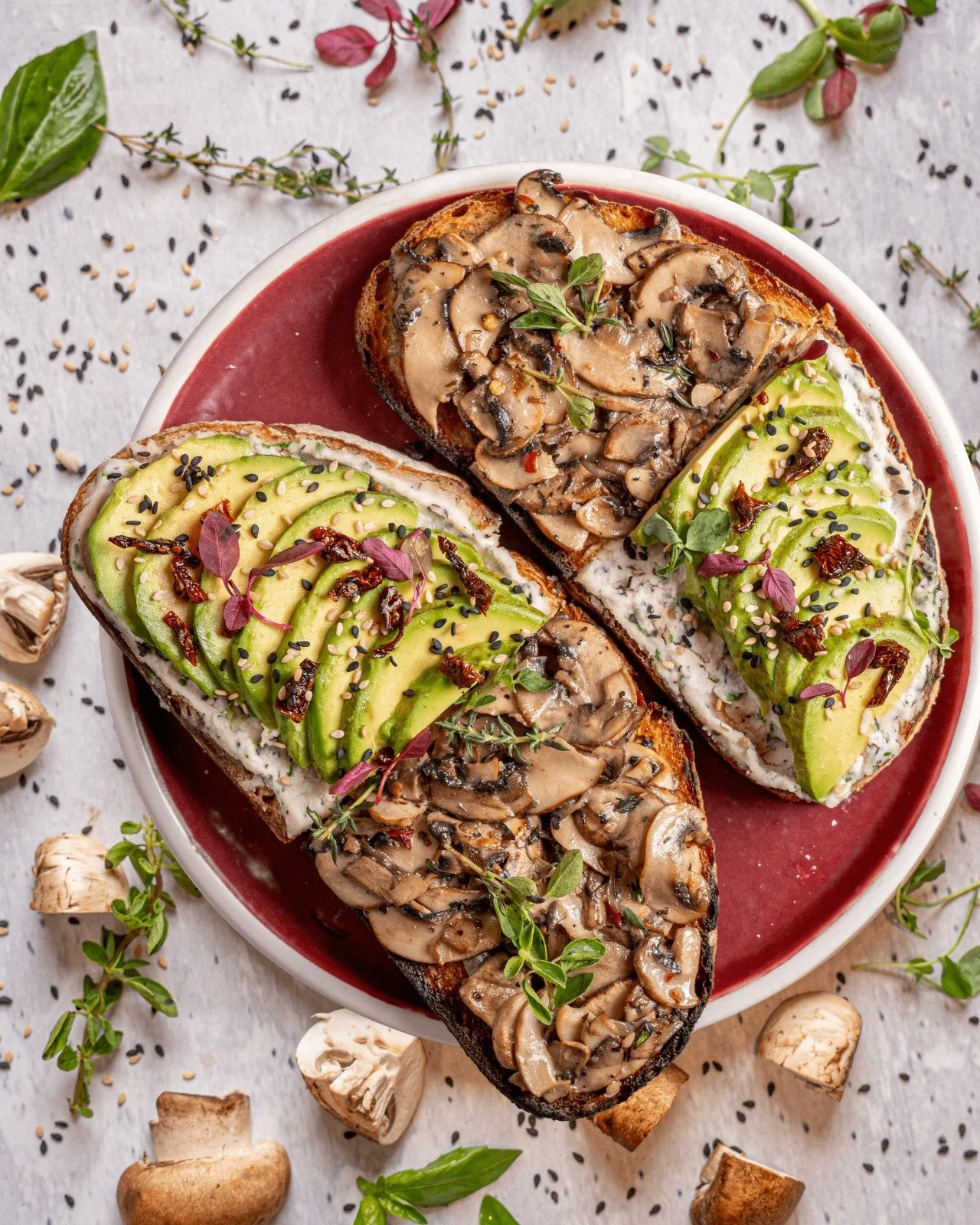 Open-faced mushroom and avocado toasts garnished with herbs and seeds on a red plate, surrounded by scattered mushrooms, herbs, and seeds.