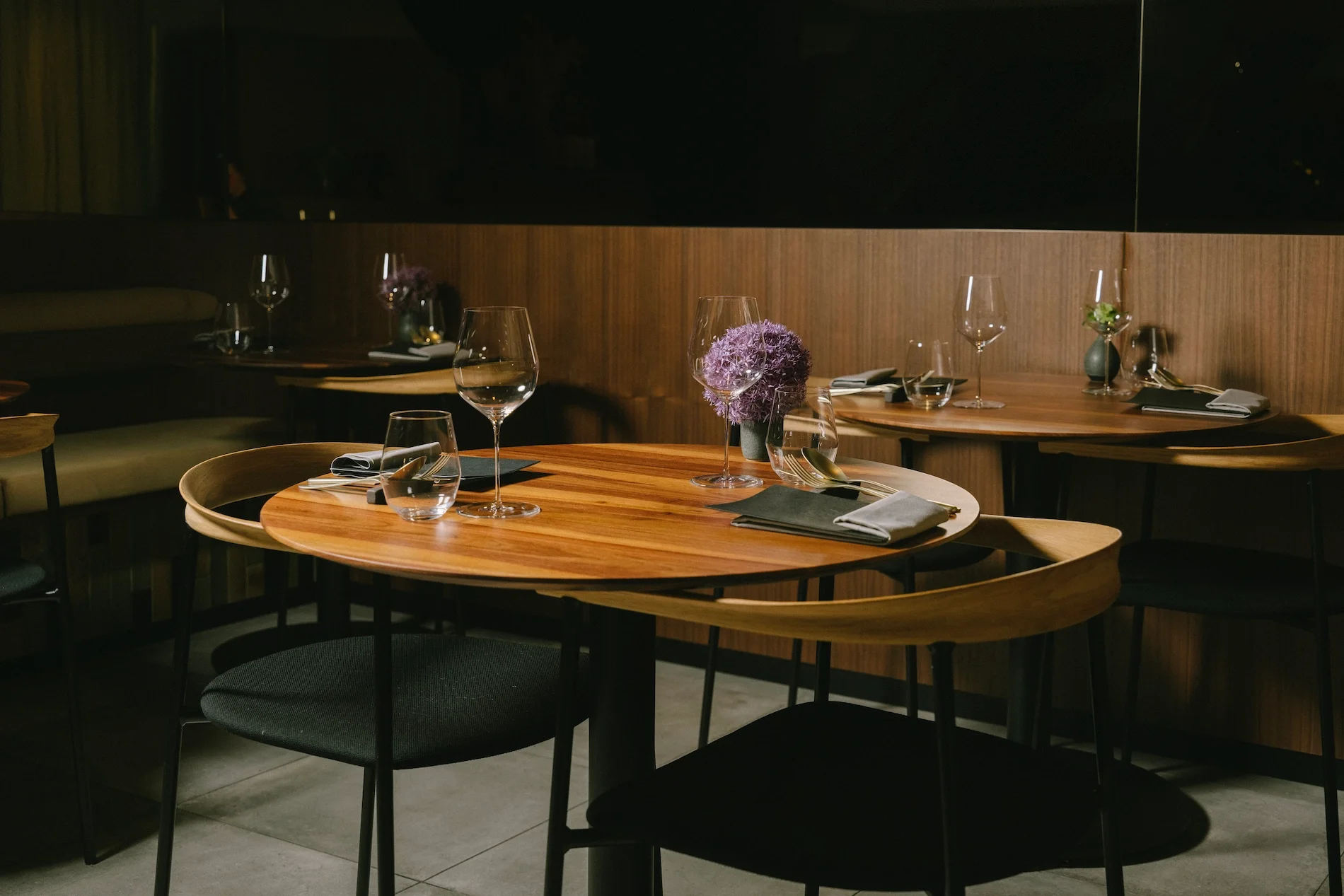 Elegant restaurant dining area with round wooden tables, black chairs, wine glasses, and cloth napkins, decorated with purple and green floral arrangements in vases.