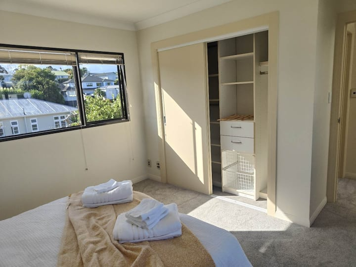 A bedroom with a bed, folded towels, a window showing a suburban neighborhood, and an open closet without doors.
