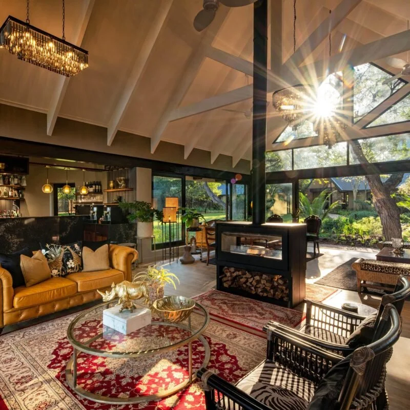 Living room with a yellow leather sofa, a glass coffee table, and a fireplace, with large windows showing a garden outside and sunlight shining through.