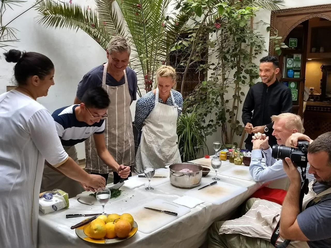 People gathered around a table in a kitchen, smiling and laughing during a cooking class. Two women are actively preparing food while others look on cheerfully. There are various ingredients and kitchen tools on the table.