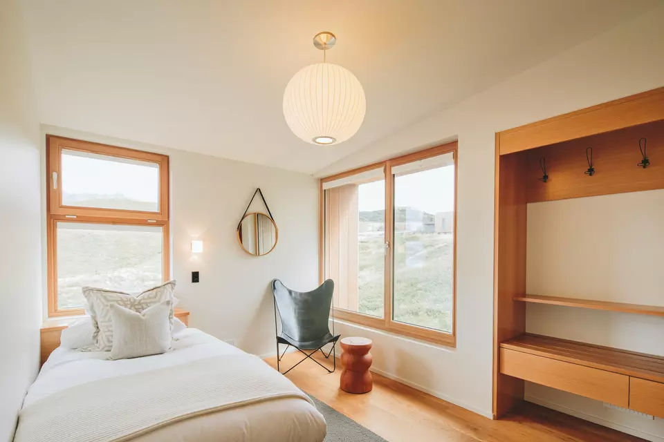 A cozy bedroom with wooden trim, a bed with white bedding and pillows, a black wingback chair, a round mirror on the wall, wooden shelving with hooks, and large windows revealing a grassy landscape outside.