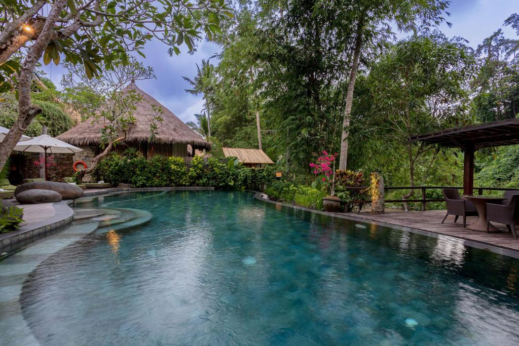 A tropical poolside scene with lush green trees, pink flowers, a thatched-roof hut, and a shaded seating area with chairs and a table.
