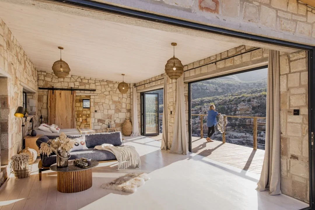 Living room with stone walls, large windows, and sliding glass doors opening to a balcony with a woman looking out at a canyon landscape.