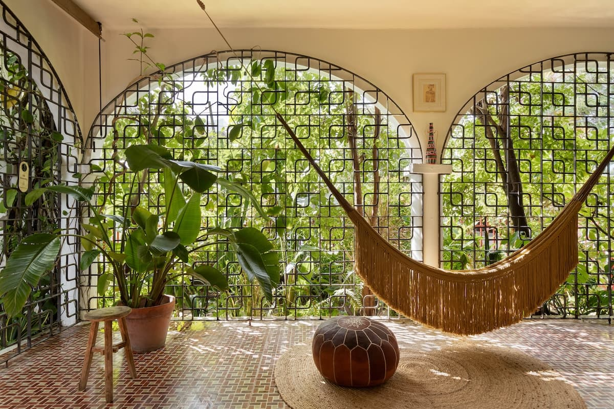 A cozy indoor space with large green plants, a hammock with brown fringe, a small wooden stool, a round leather pouf, and a natural fiber rug. The space features decorative black metal grid walls and a view of lush trees outside.