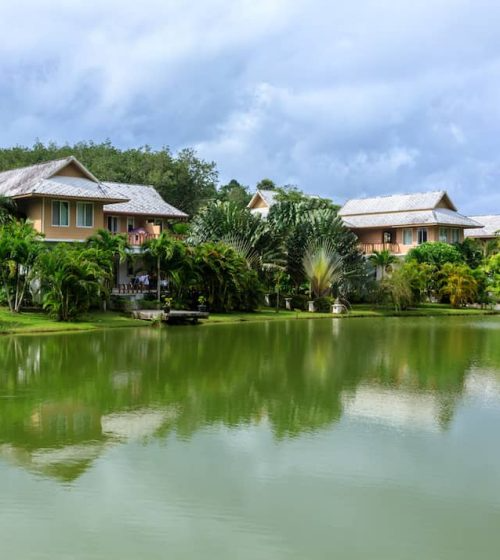 Houses with traditional rooftops by a lake surrounded by palm trees and lush greenery under a cloudy sky.