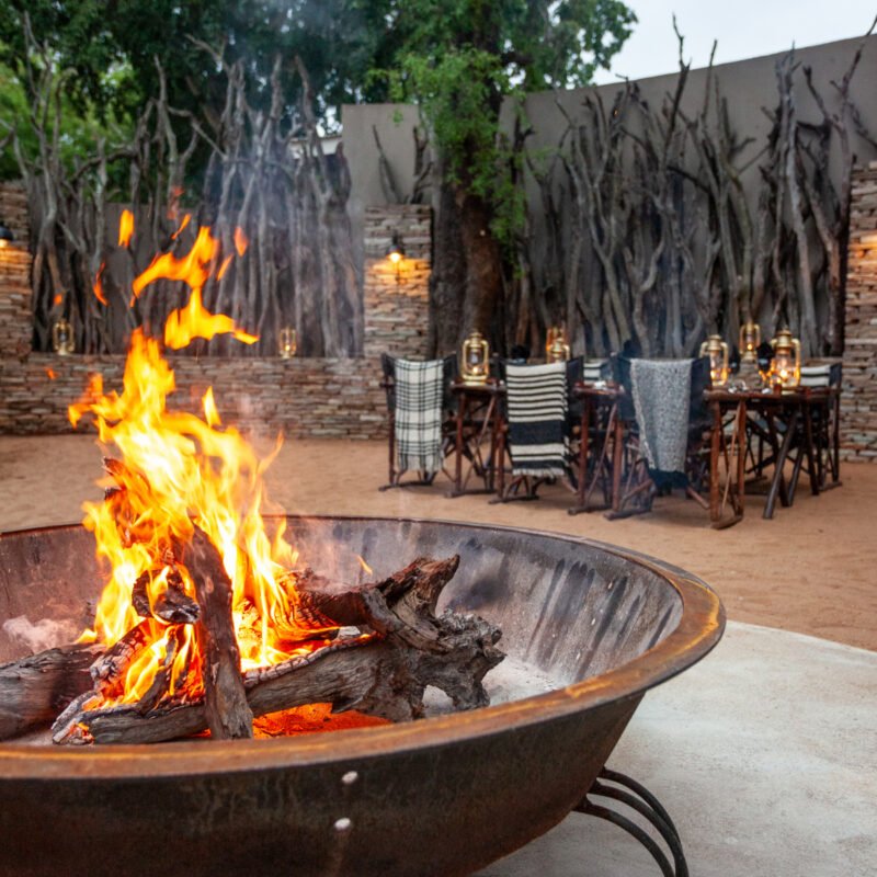 Outdoor fire pit with burning logs in the foreground, and a dining table with napkins and lanterns in the background, set against a decorative wood and stone wall.