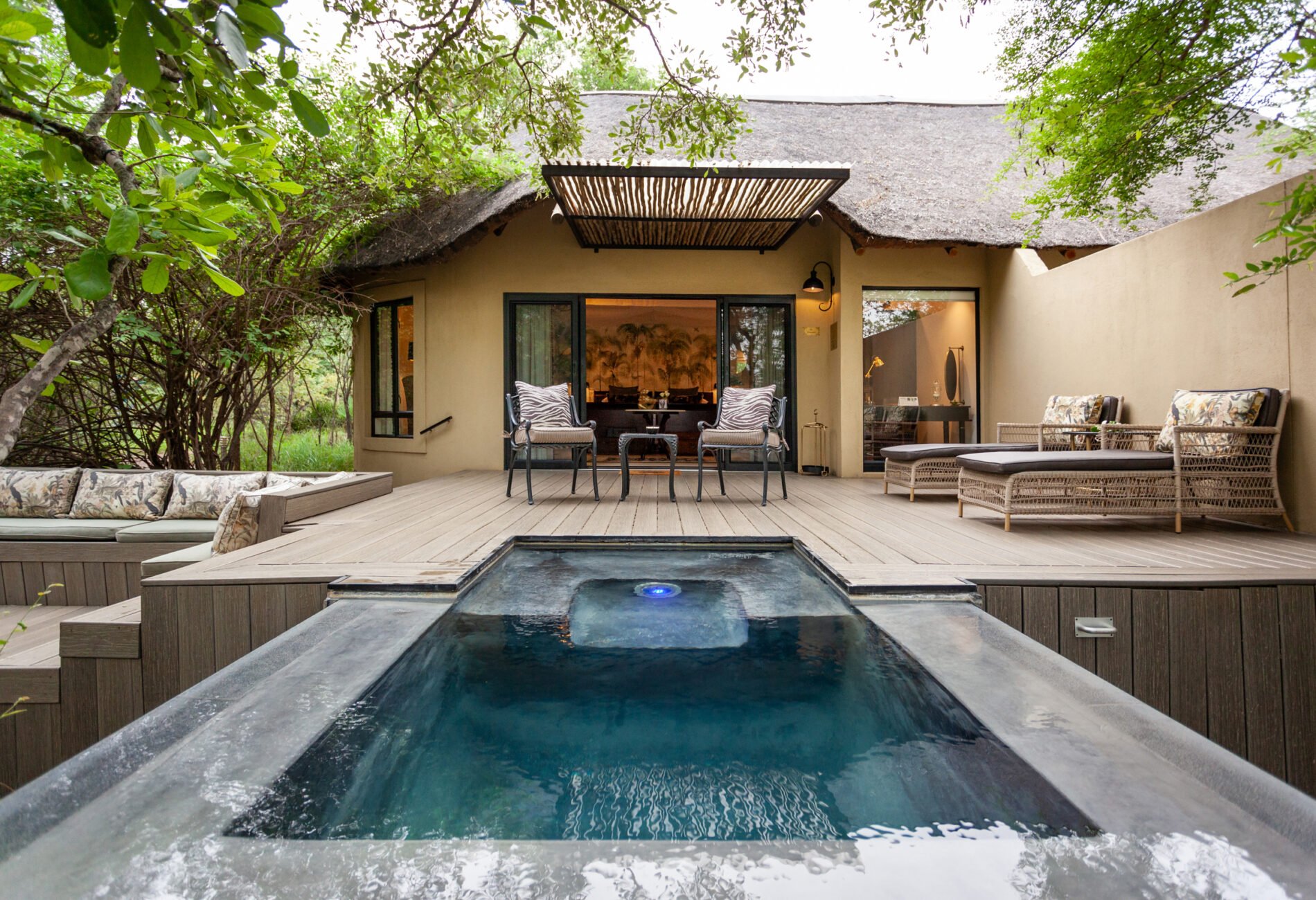 A backyard patio with a hot tub in the foreground, outdoor seating including armchairs and a bench, a two-story house with a thatched roof, surrounded by trees and greenery.