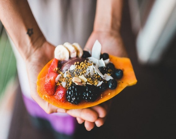 Person holding a carved papaya filled with assorted berries, bananas, and decorative toppings.