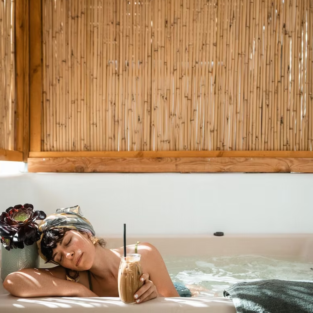 Woman relaxing in a bathtub with a beverage and a towel, surrounded by bamboo fencing.