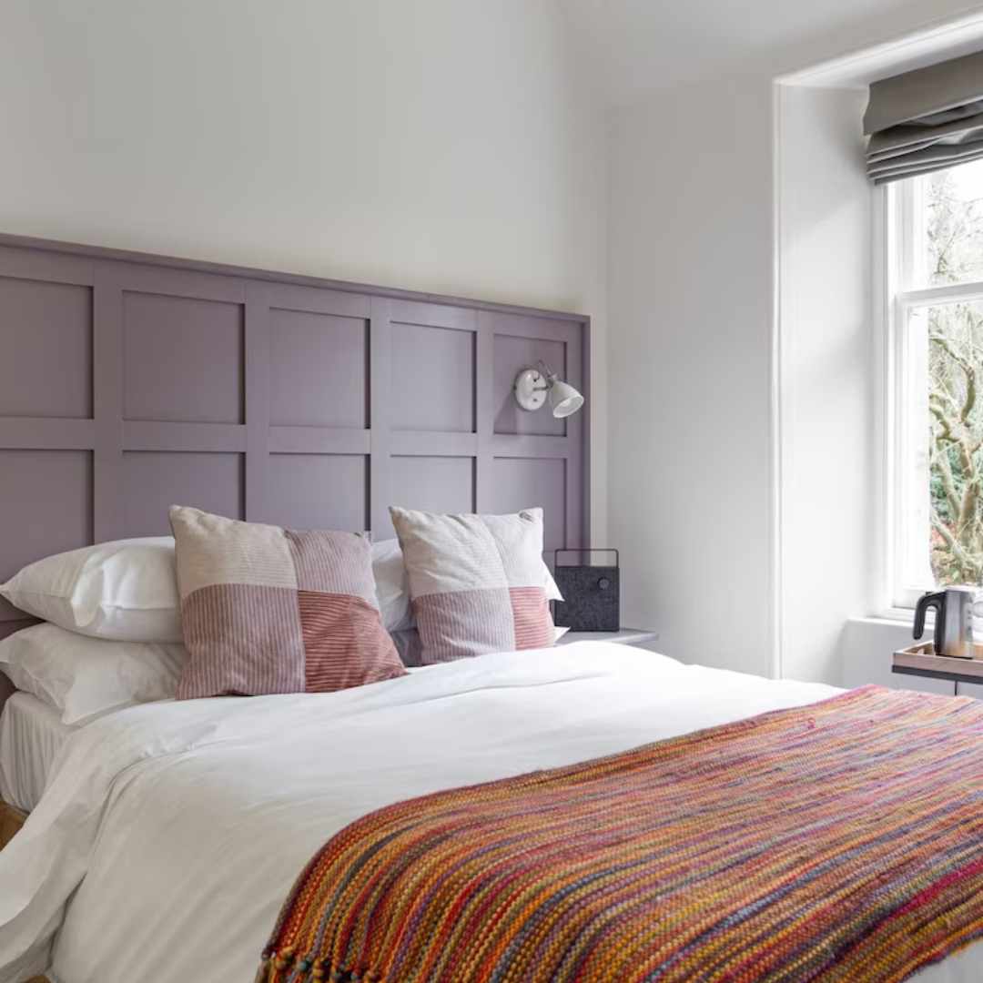 A cozy bedroom with a bed covered in white bedding, two patterned pillows, and a multicolored woven blanket at the foot. There is a lavender headboard, a reading wall lamp, a black bedside speaker, and a window with gray blinds showing trees outside.