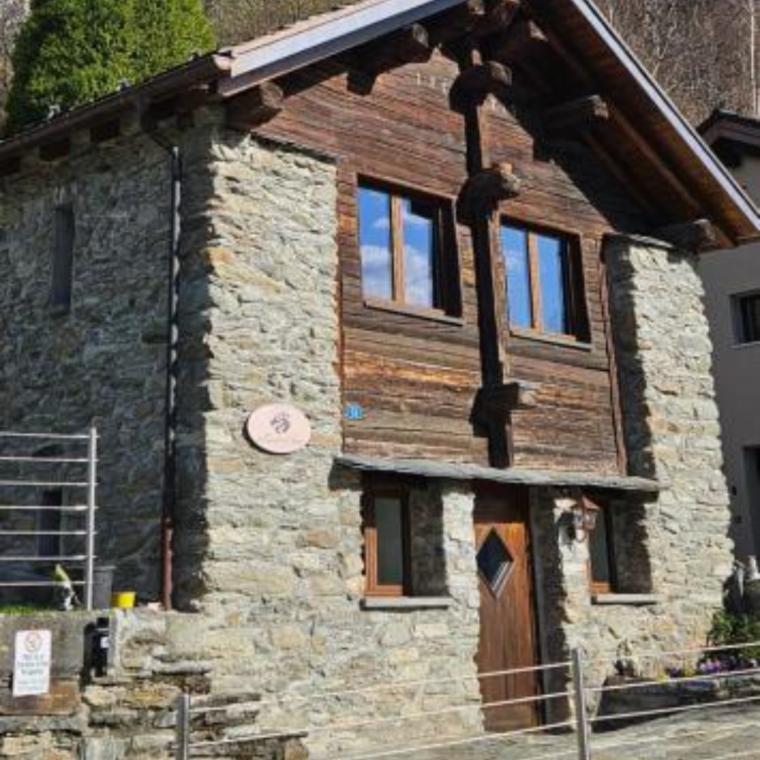 Stone and wood mountain house with a steep roof and two large windows reflecting blue sky, surrounded by a small fence and greenery.