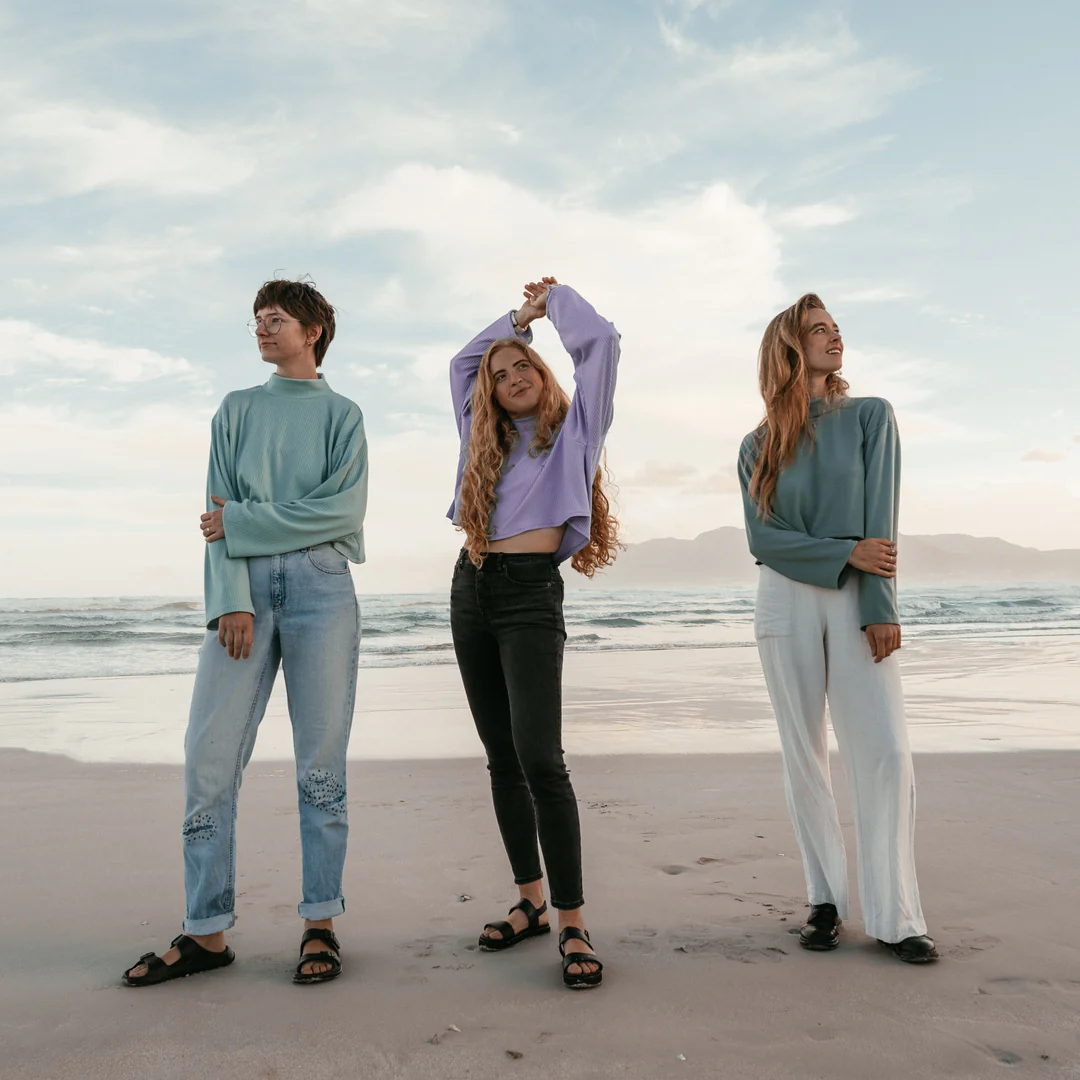 Three young women standing on the beach during sunset, dressed casually, with ocean waves and a cloudy sky in the background.