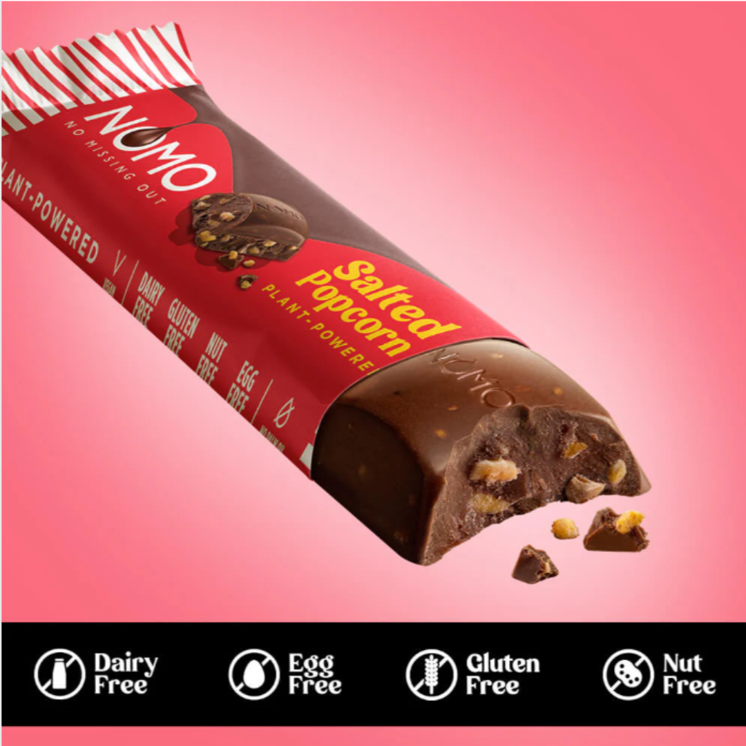 A bar of dairy-free, egg-free, gluten-free, nut-free layered dark chocolate with nuts, lying on a pink background with a bite taken out of it. The bar is partly wrapped in red packaging labeled "Sweetened Popcorn plant-powered".