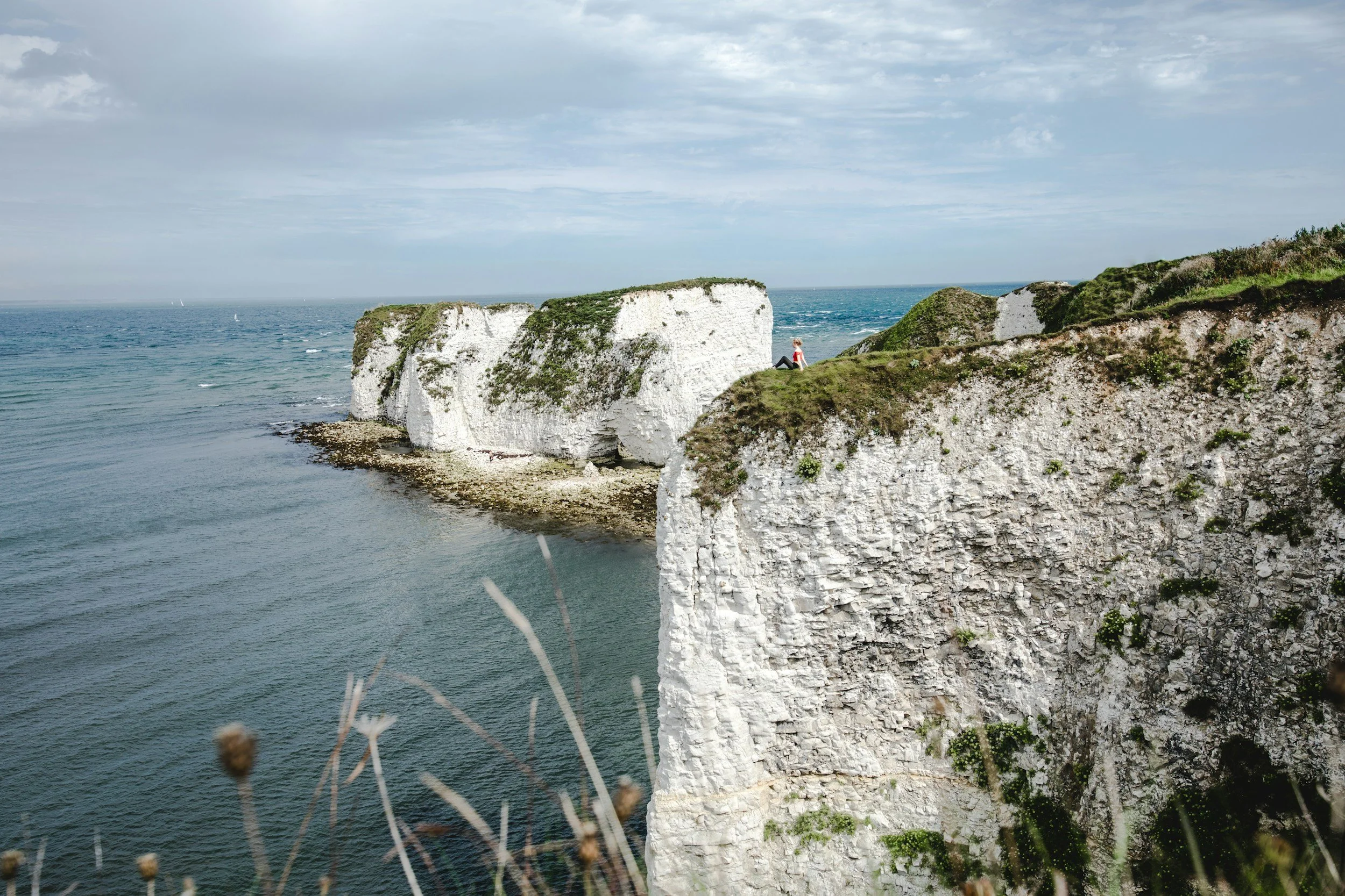 Cliffs along the coast with a person sitting on the edge, overlooking the sea. The sky is cloudy and there are sailboats in the distance.