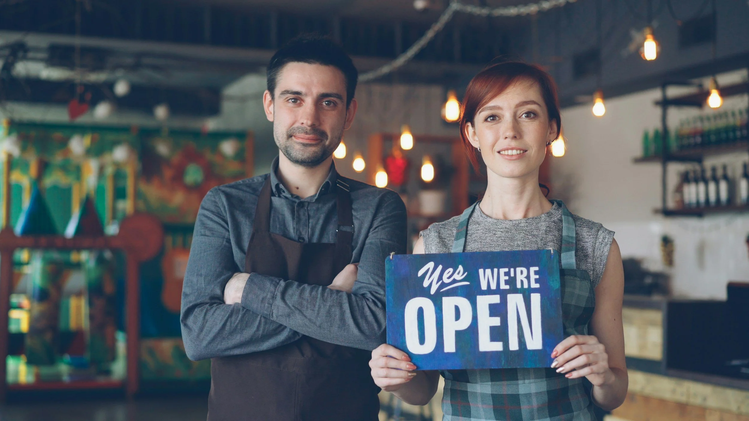 A man and a woman standing inside a store, both wearing gray aprons. The woman is holding a sign that reads "Yes, we're open." The background shows shelves with products and hanging lights.