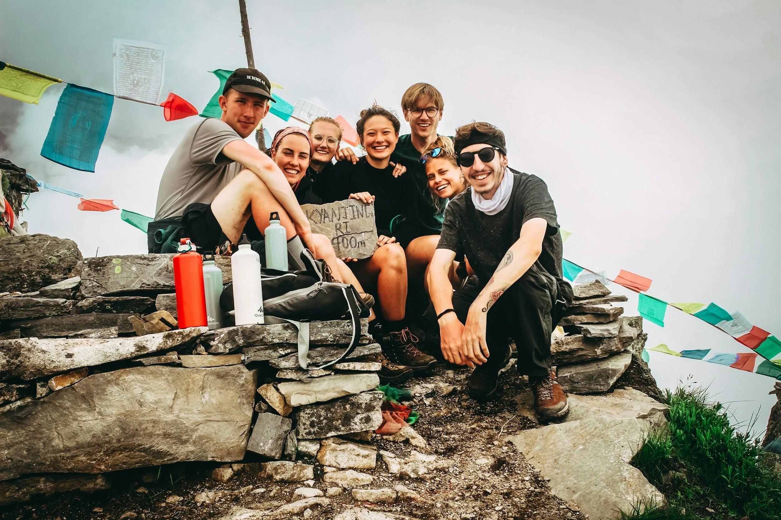 Group of eight hikers sitting on a rocky summit with prayer flags in the background, smiling and holding a sign that reads "Kyanjing Ri 4000m," with water bottles and backpacks nearby.
