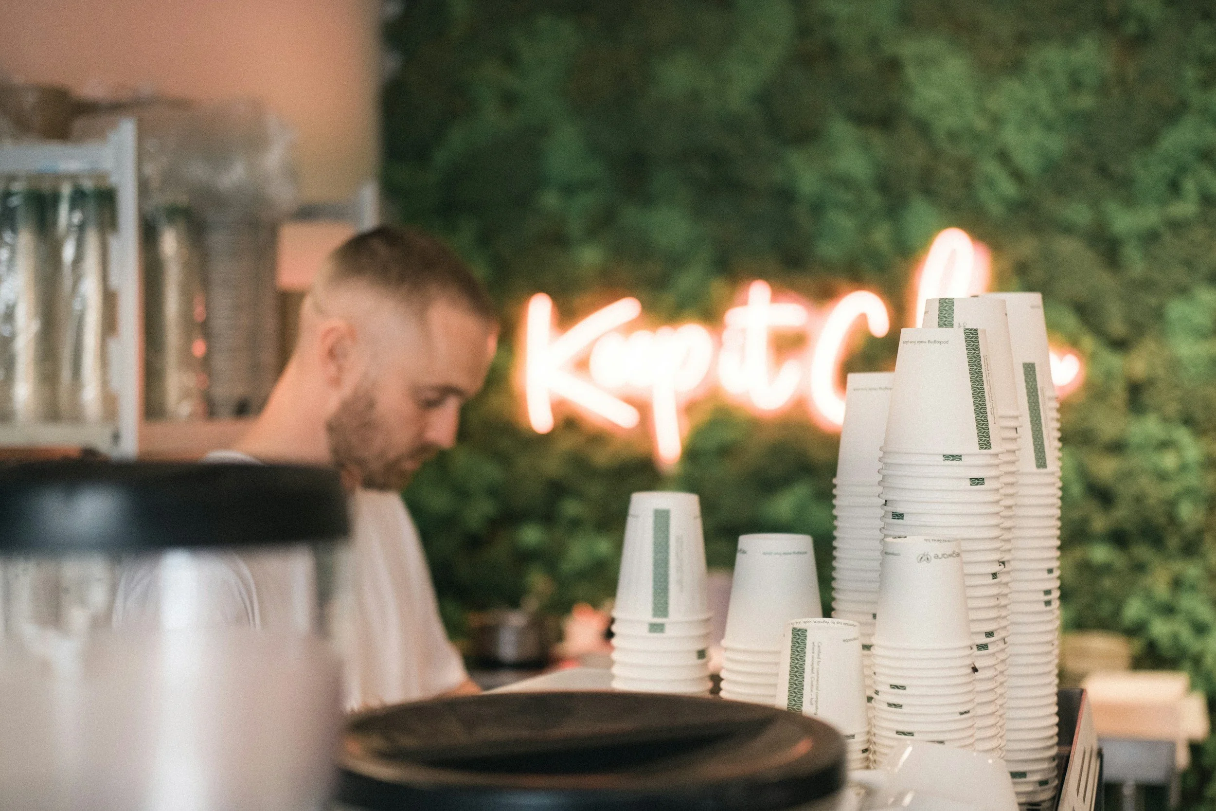 A person working behind a counter with a stack of paper cups in front of them, and a neon sign with the text "Kp Tea" in the background.