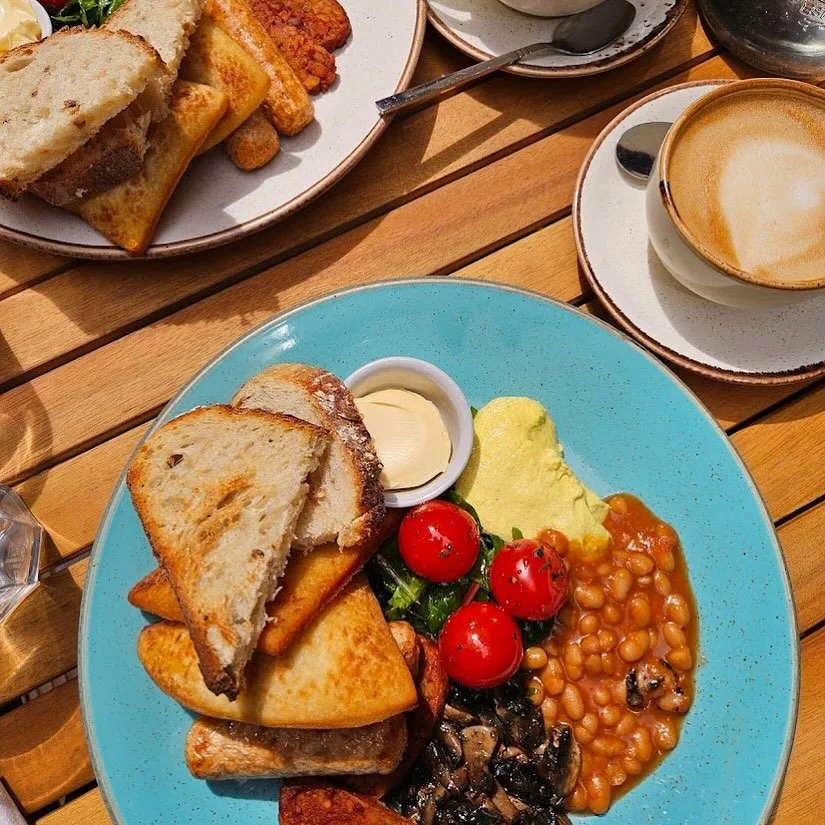 Full English breakfast with baked beans, potatoes, tomatoes, mushrooms, scrambled eggs, toast, and butter, served with a cappuccino on a wooden table.