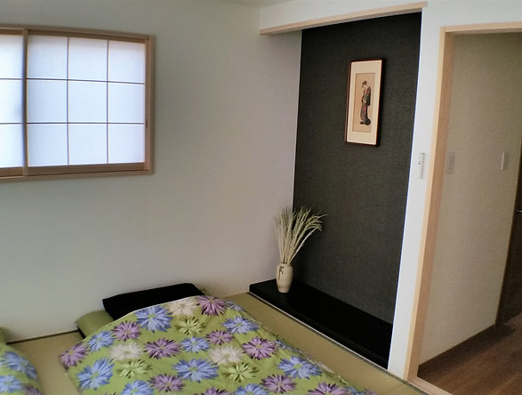 A bedroom with a bed featuring a floral bedspread, a window with paper shoji-style blinds, a black accent wall with a framed art piece, a potted plant on a shelf, and an open doorway leading to another room.