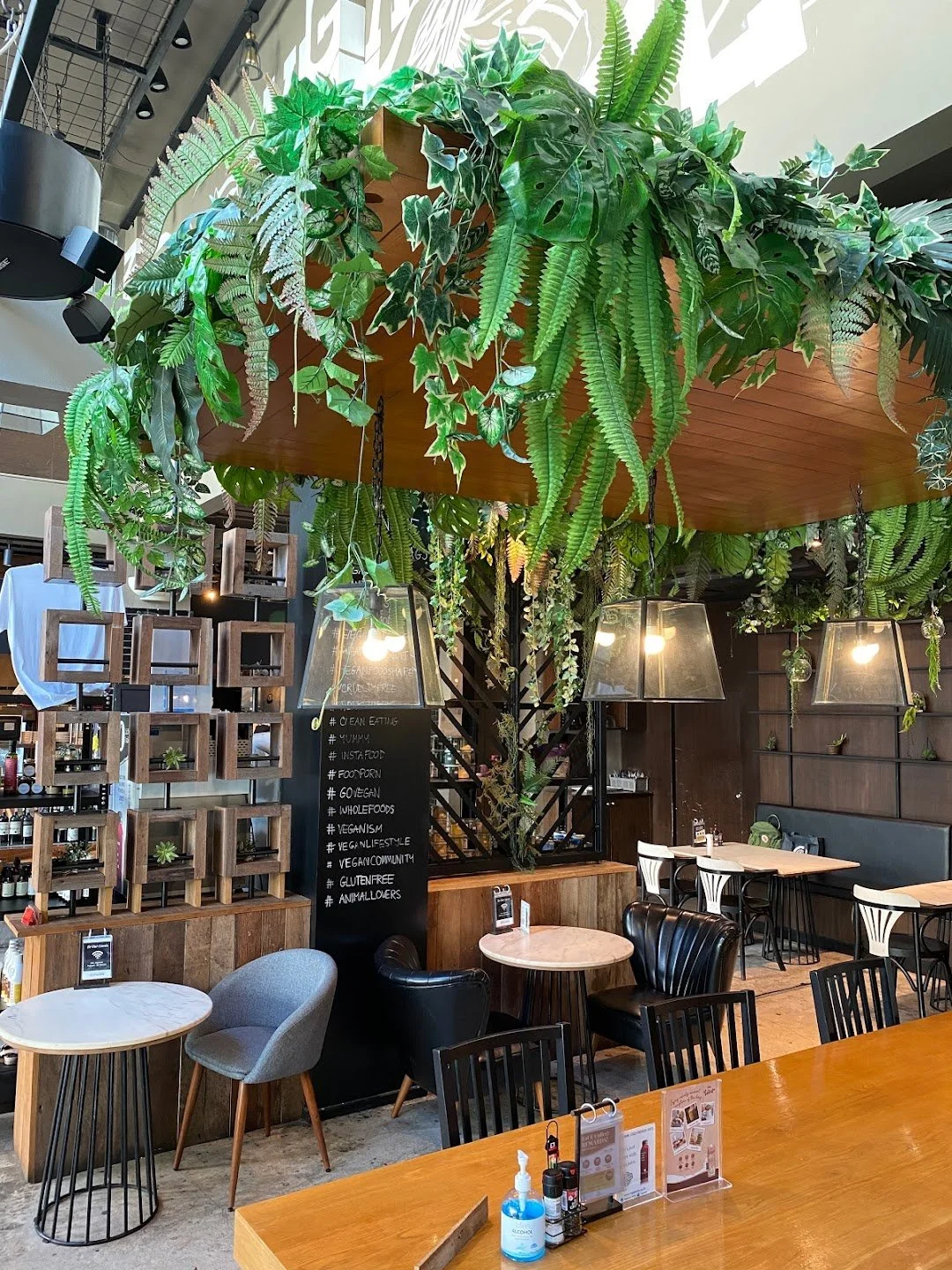 Indoor restaurant with hanging plants and greenery overhead, wooden furniture, black and white chairs, and a black chalkboard with handwritten menu.
