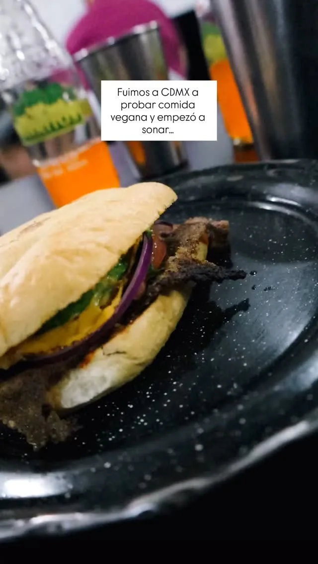 A vegan burger in a bun with lettuce, tomato, red onion, and a black patty, placed on a black plate with a slightly blurred background of cups and a pink object. A text overlay in Spanish mentions traveling to Mexico City to try vegan food.