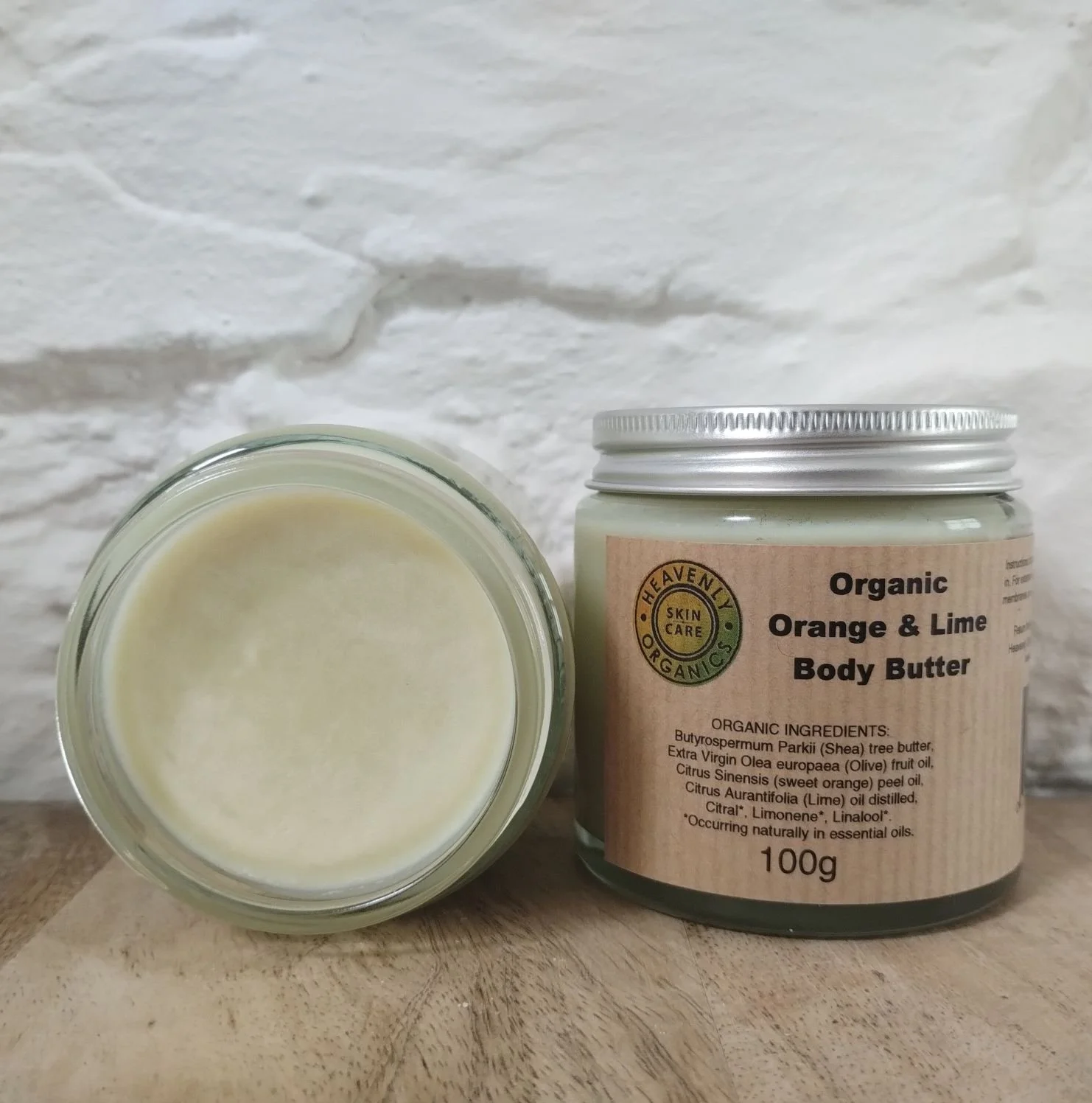 A jar of organic orange and lime body butter with a label and a small bowl of the body butter in front of it, placed on a wooden surface against a white textured wall.