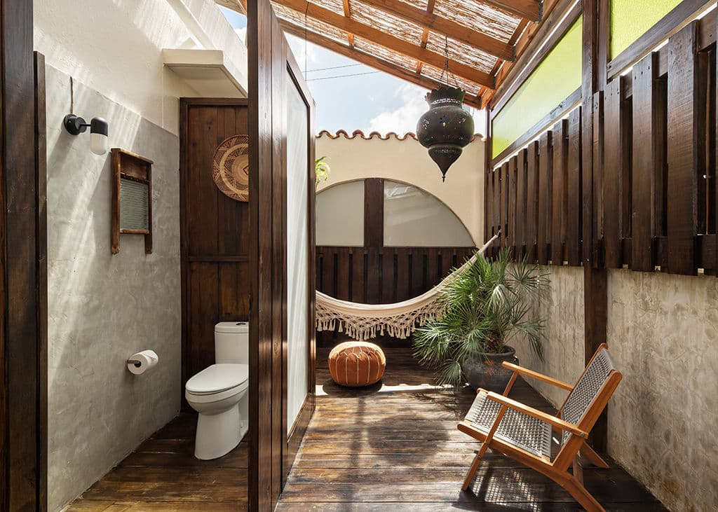 An outdoor bathroom with a toilet, hammock, and seating area with a wooden chair. Wooden and concrete walls, a potted plant, and a hanging lantern.
