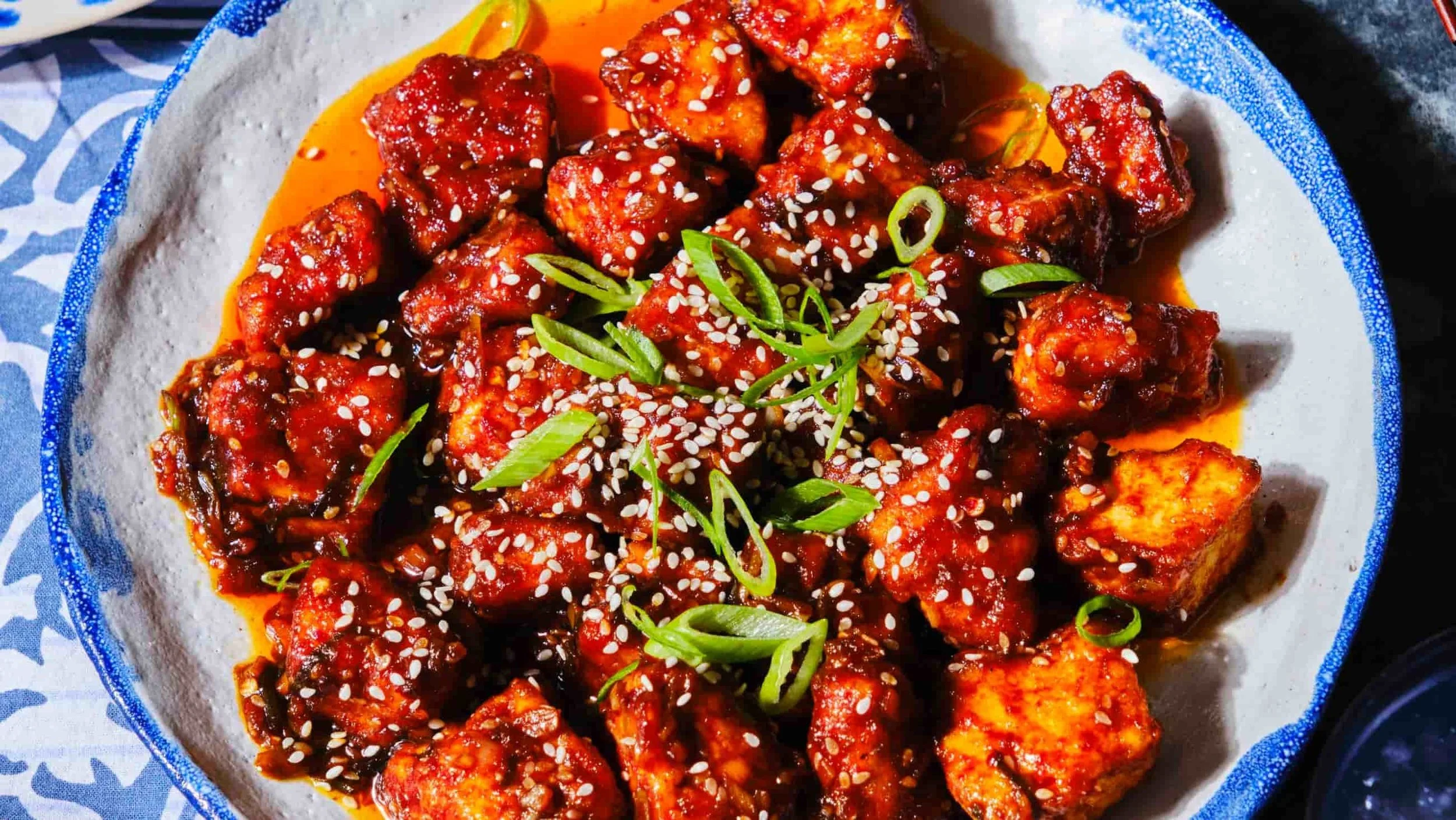 Plate of spicy Asian-style chicken with sesame seeds and green onions, served on a ceramic dish.