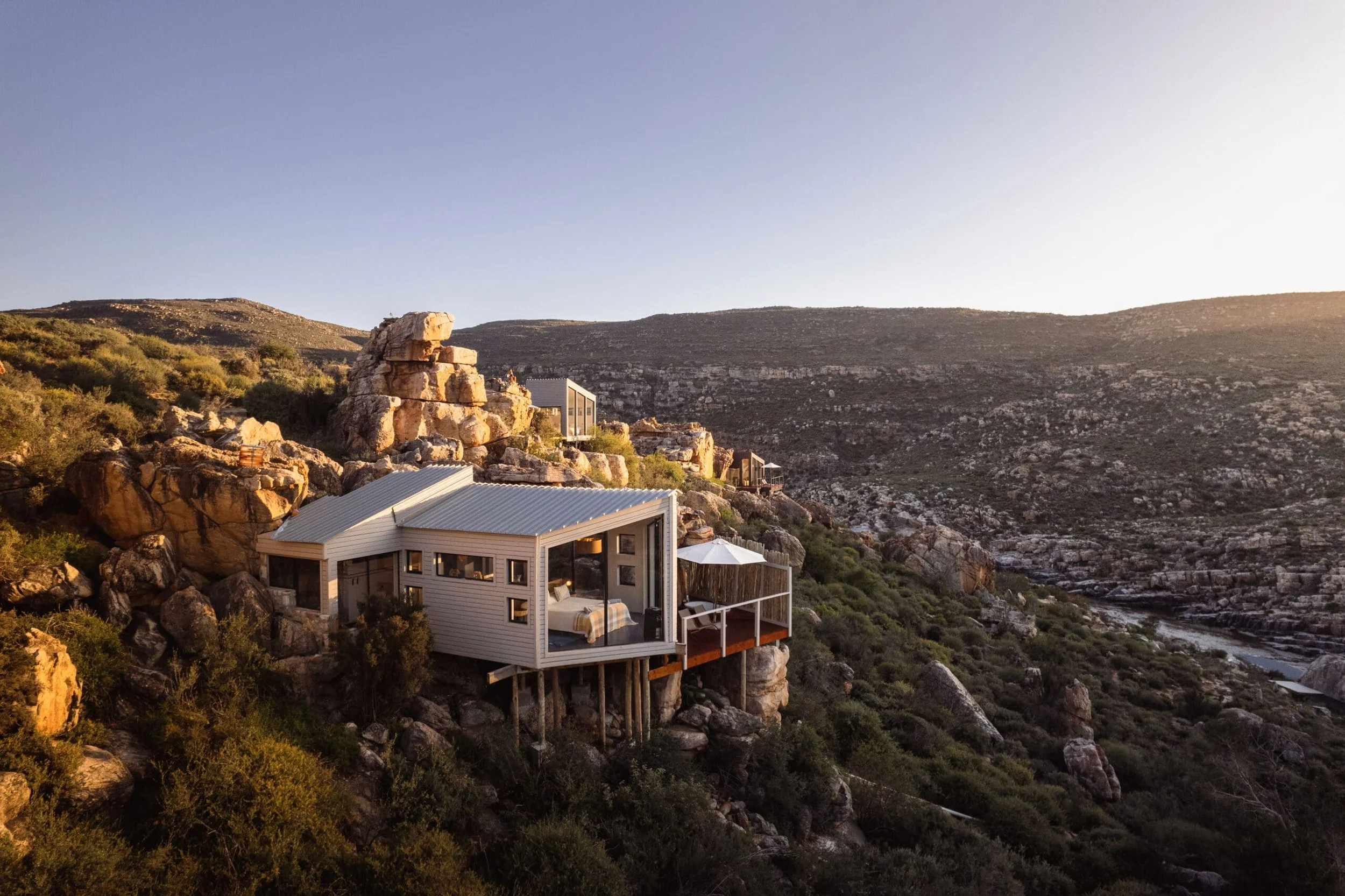 A modern white house built on stilts among large rocks on a hillside, overlooking a dry, mountainous landscape during sunset.