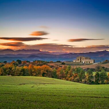 Landscape with a green field in the foreground, colorful trees, a building on a hill, mountains in the background, and a partly cloudy sunset sky.