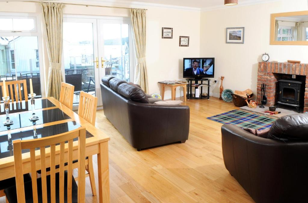 Open-plan living room and dining area with wooden floors, a dining table with chairs, a sofa, an armchair, a brick fireplace with tools and a plaid rug, a TV on a stand, and large glass doors leading to a balcony.