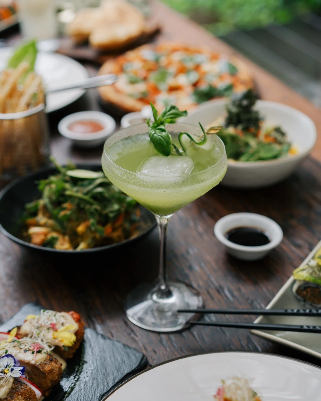 A table set with various Asian dishes including a green cocktail with a basil garnish in the foreground, bowls of salad, sushi, pizza, and soy sauce alongside black chopsticks.