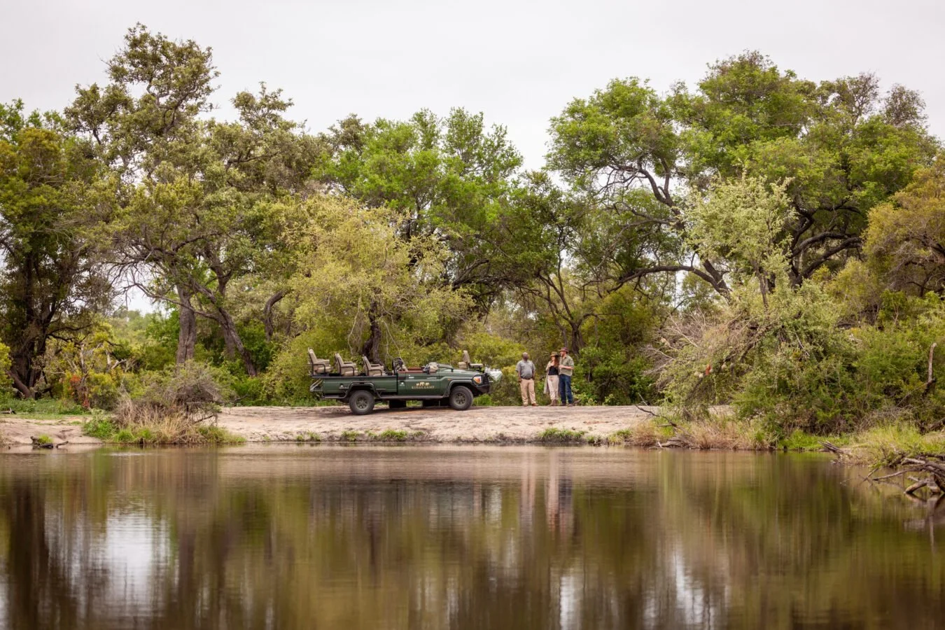 Group of people talking next to a safari vehicle near a riverbank surrounded by trees.