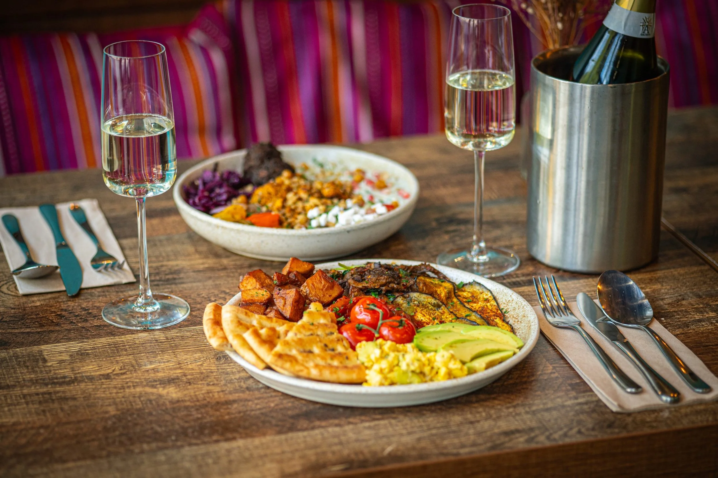 A dinner table with two glasses of white wine, a bowl of rice, a pizza with cherry tomatoes and avocado slices, and a plate of various grilled and cooked foods, all set on a wooden table with cutlery.