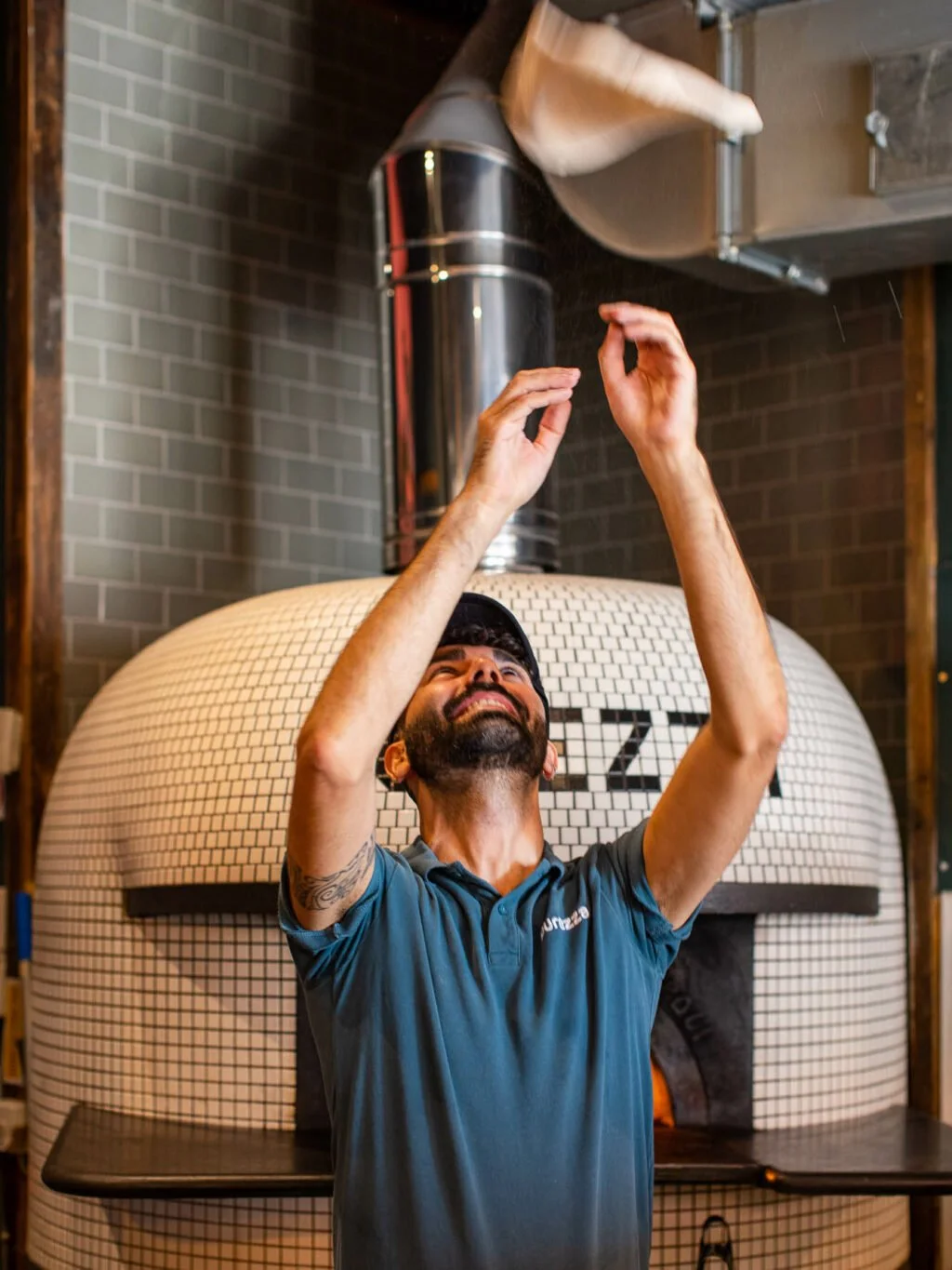 A man smiling and raising his arms in front of a wood-fired pizza oven in a pizzeria.