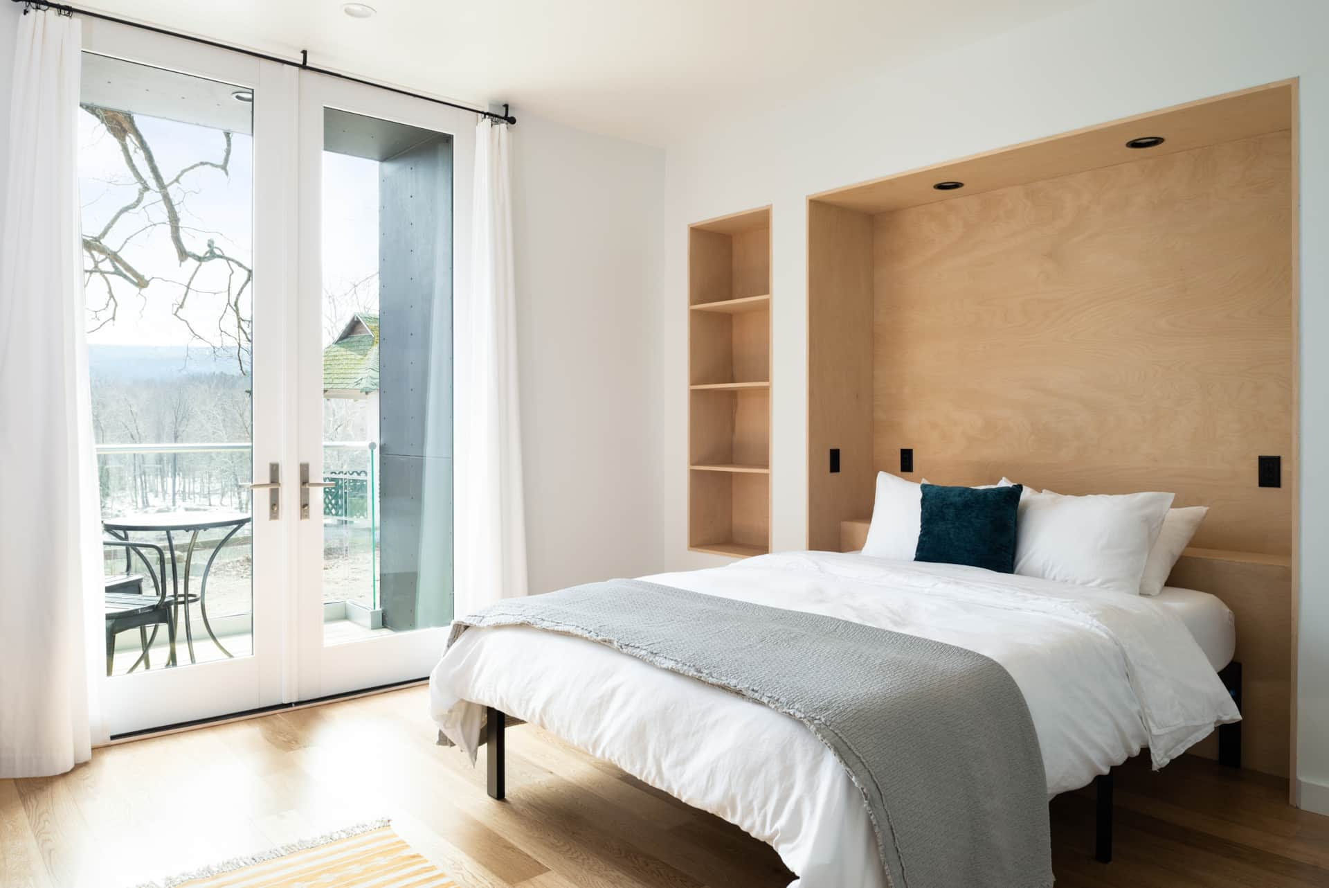 A modern bedroom with white walls, a bed with white sheets and pillows, a wooden headboard with built-in shelves, and a sliding glass door opening to a balcony with a table and chairs, overlooking a scenic outdoor landscape.