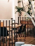 Cozy cafe interior with wooden tables and black chairs, plant decor, and large windows letting in natural light.