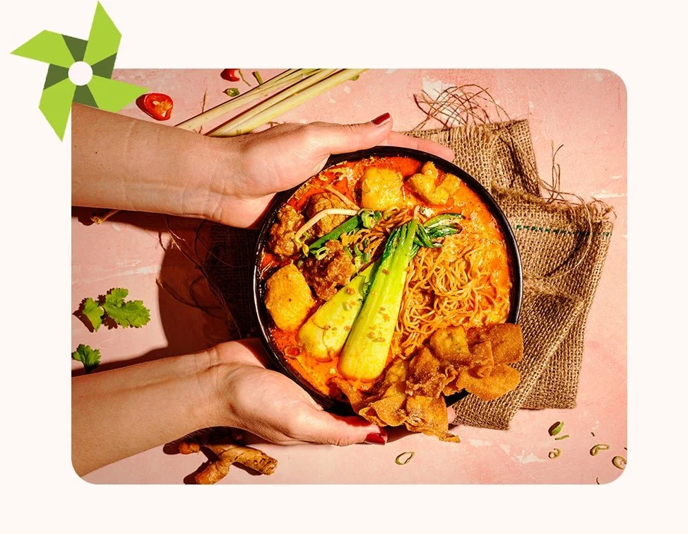 A bowl of spicy ramen with vegetables and fried toppings, held by two hands over a pink surface with garnish, leek, and shredded bamboo on the side.
