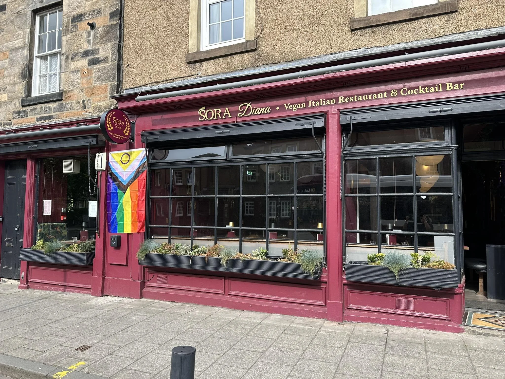 The exterior of a vegan Italian restaurant and cocktail bar called Sora Diana. The storefront has large glass windows, a maroon-colored facade, and a rainbow pride flag hanging outside.
