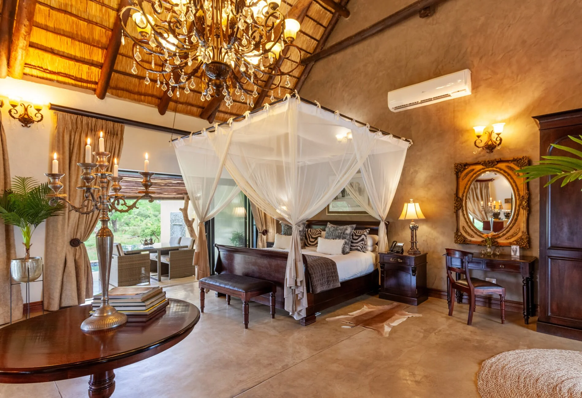Luxury bedroom with a four-poster bed, chandelier, air conditioner, and ornate mirror, with warm lighting and tropical decor.