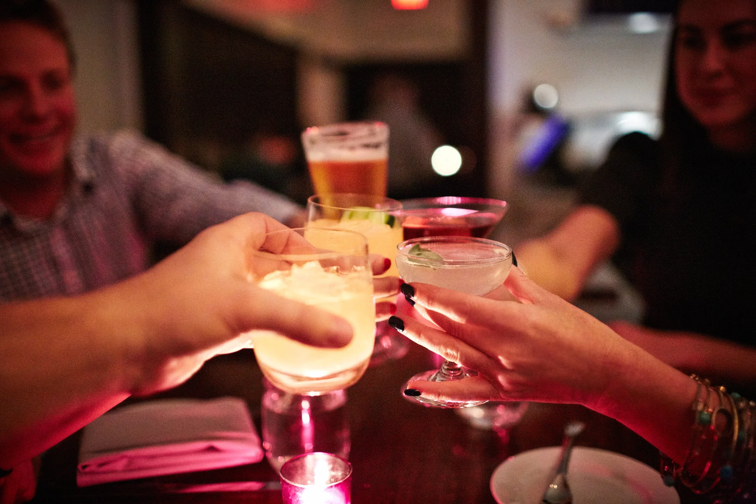 People raising cocktail glasses for a toast in a dimly lit setting, likely a bar or nightclub.