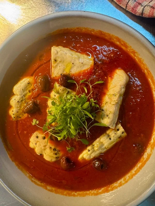 A bowl of tomato-based soup with pieces of fish, black olives, and garnished with fresh herbs.
