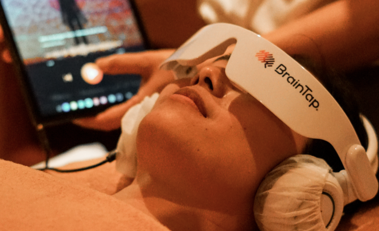 Person wearing a BrainTap headset lying down with their eyes closed, with a tablet or monitor displaying a blurred image in the background.